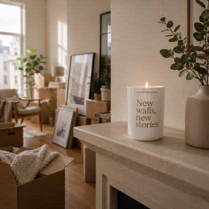 A white scented candle with gold lettering reading 'New walls, new stories' sits lit on a light stone mantel in a softly lit, warm-toned room with framed art, plants, and moving boxes in the background.