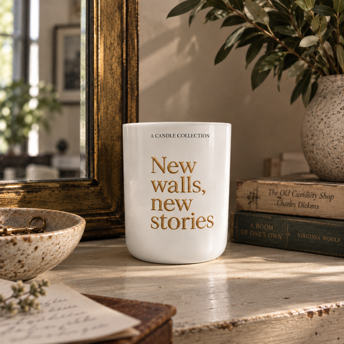 New walls, new stories candle by A Candle Collection, white vessel with gold text, styled on a sunlit shelf with books and a plant